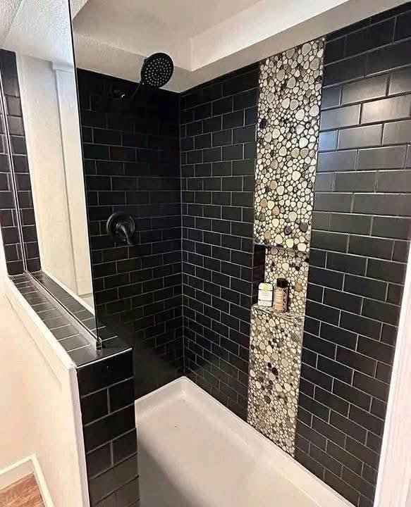 Modern shower with black rectangular tiles and a pebble accent strip. Features a rainfall showerhead and built-in shelf holding toiletries.