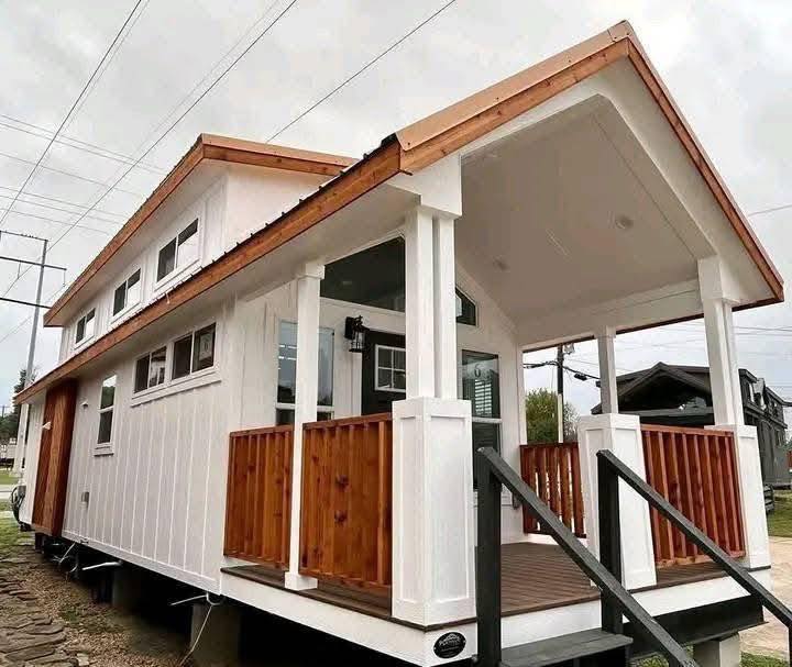 A modern tiny house with a white exterior and wood accents. It features a cozy front porch and black stairs, creating a welcoming and compact design.