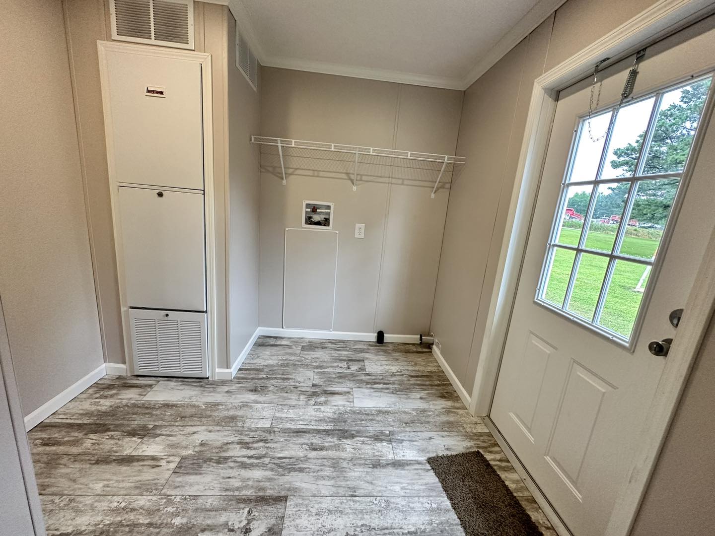 A small laundry room with beige walls and wood-textured flooring. It features a white utility cabinet, a wire shelf, and a glass-paneled door leading outside. The scene is calm and tidy.