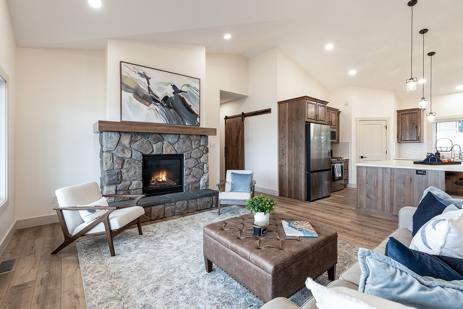 Bright living room with stone fireplace, abstract art, and leather ottoman. Cozy armchairs and soft lighting create a warm, inviting atmosphere.