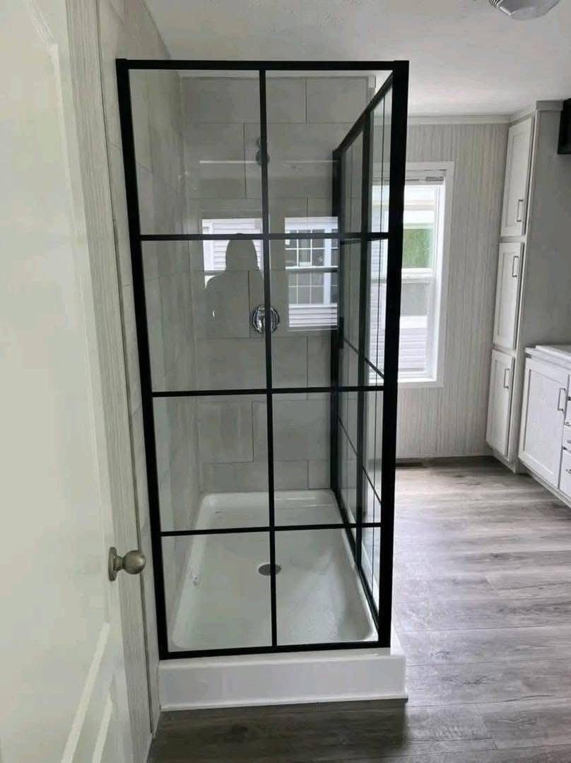 A modern bathroom features a glass shower with black grid frames, set against light gray tiles. Sunlight filters in from a window, creating a clean and airy feel.