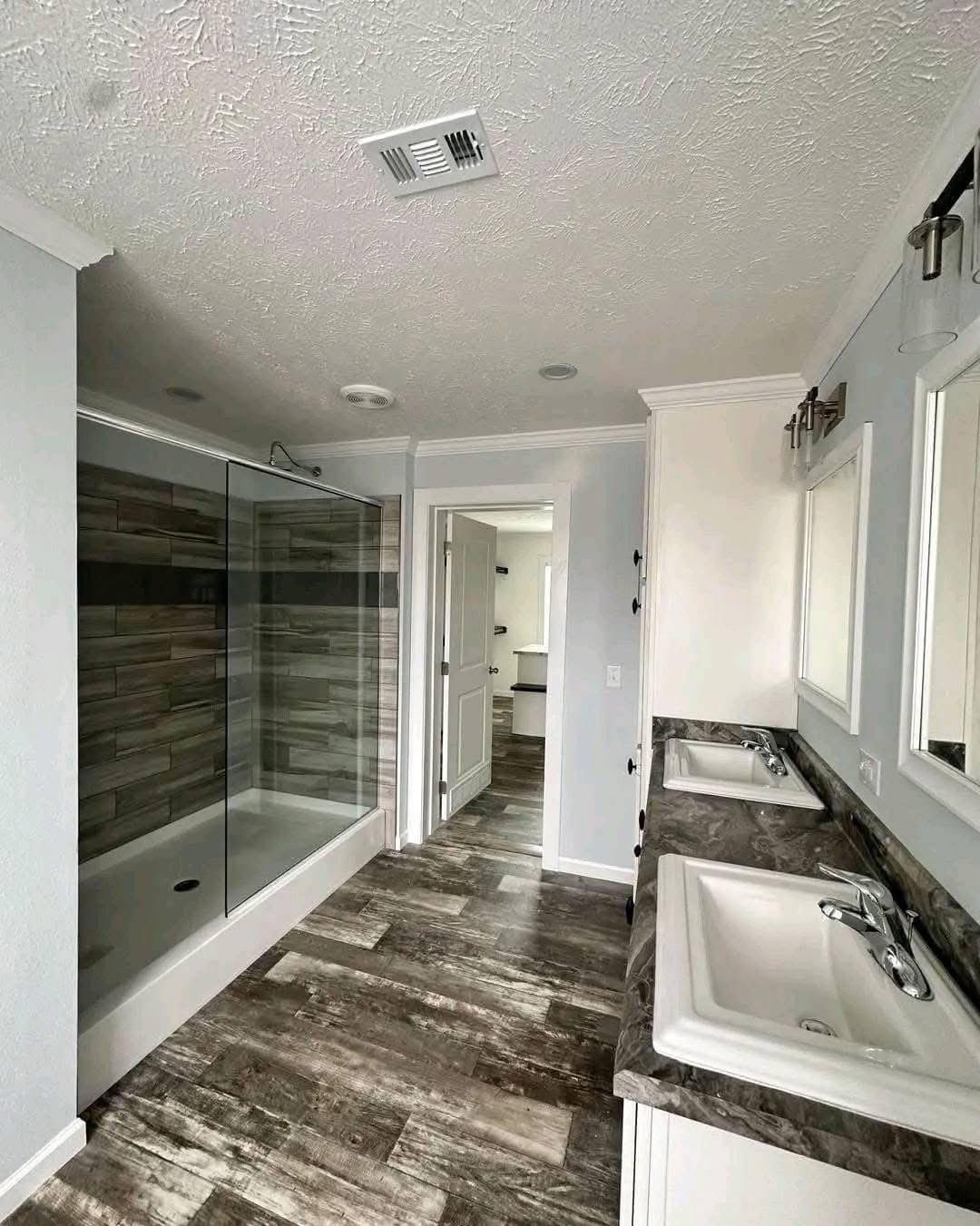 Spacious modern bathroom with a large glass shower, dual sinks with dark countertops, and a wood-patterned floor. Bright and clean atmosphere.