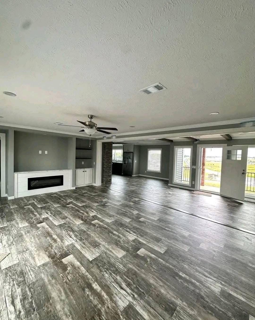 Spacious, open-concept room with rustic gray wood floors, neutral gray walls, ceiling fan, modern built-in fireplace, large windows, and glass door leading outside.