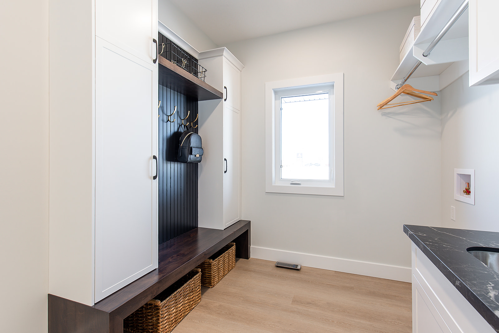 Bright, modern mudroom with white cabinetry and light wood flooring. Features coat hooks, a black bench, wicker baskets, a window, and a countertop.