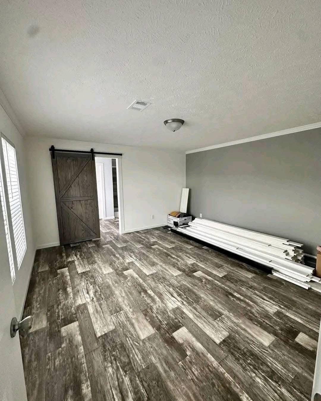 An empty room with grayish wood flooring and a gray accent wall. A stack of white trim lies on the floor, and a dark barn-style sliding door stands open.