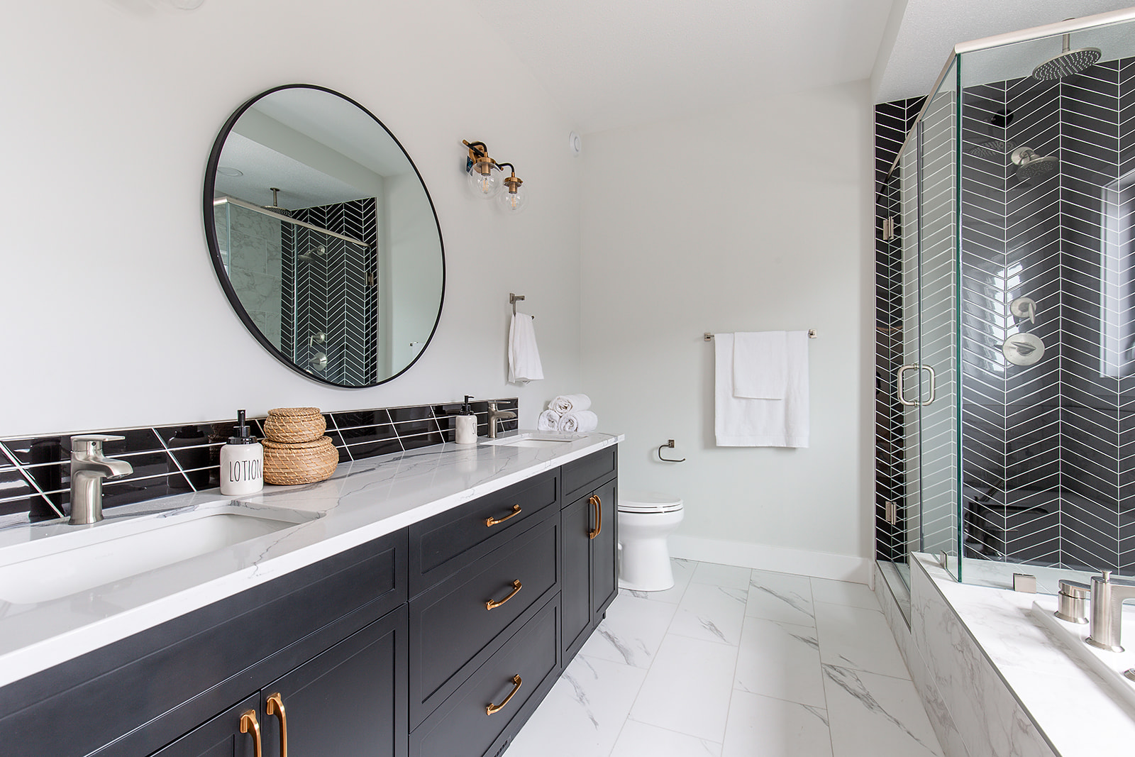 Modern bathroom with sleek design. Features a large round mirror above a black vanity with white countertop, brass handles, and minimal decor. Marble tile floor and glass shower with black herringbone tiles. Bright, clean, and elegant aesthetic.