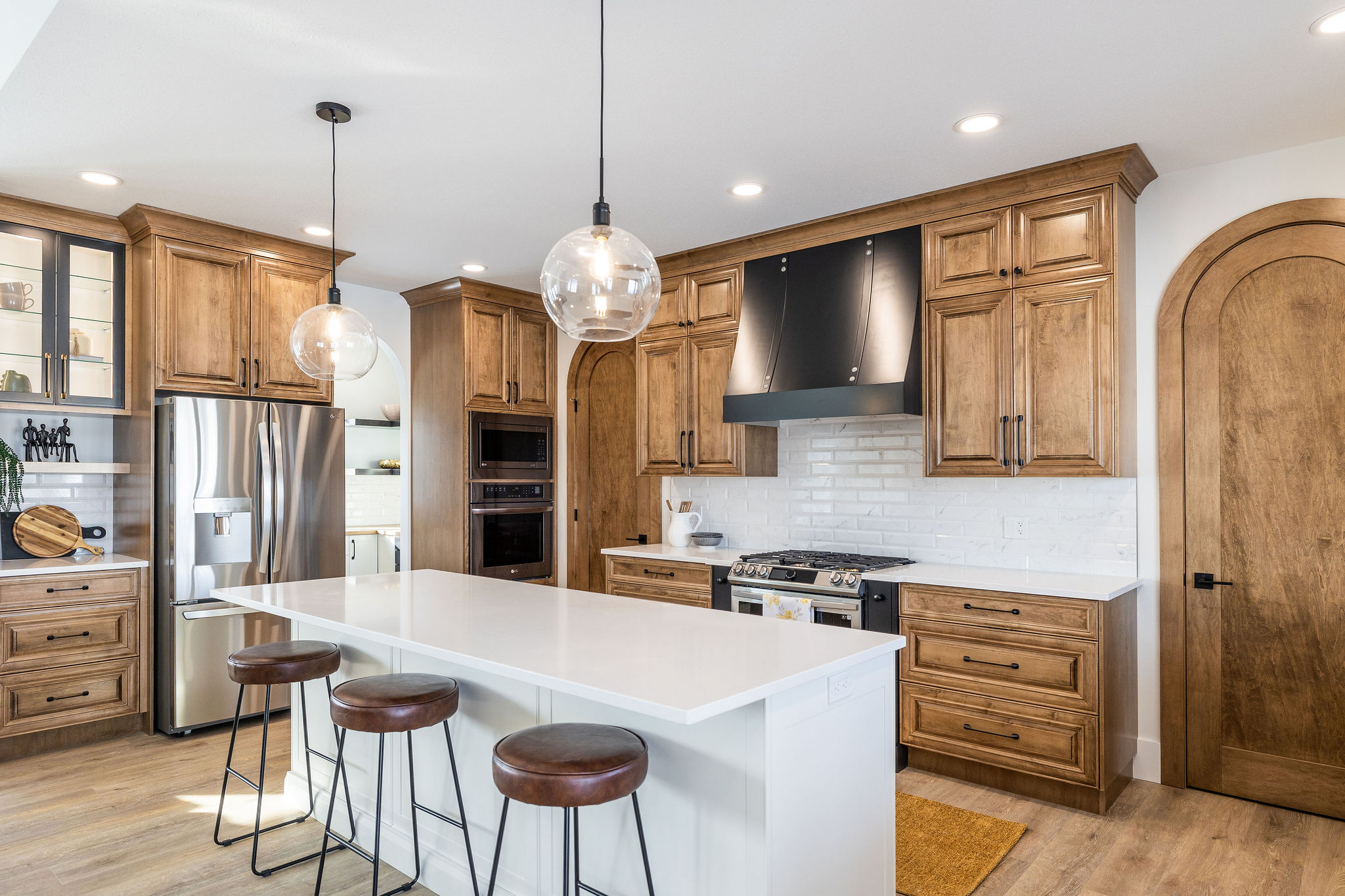 A modern kitchen with wooden cabinets, white countertops, a stainless steel fridge, and a central island with three stools. Two globe pendant lights hang above, creating a warm and inviting atmosphere.