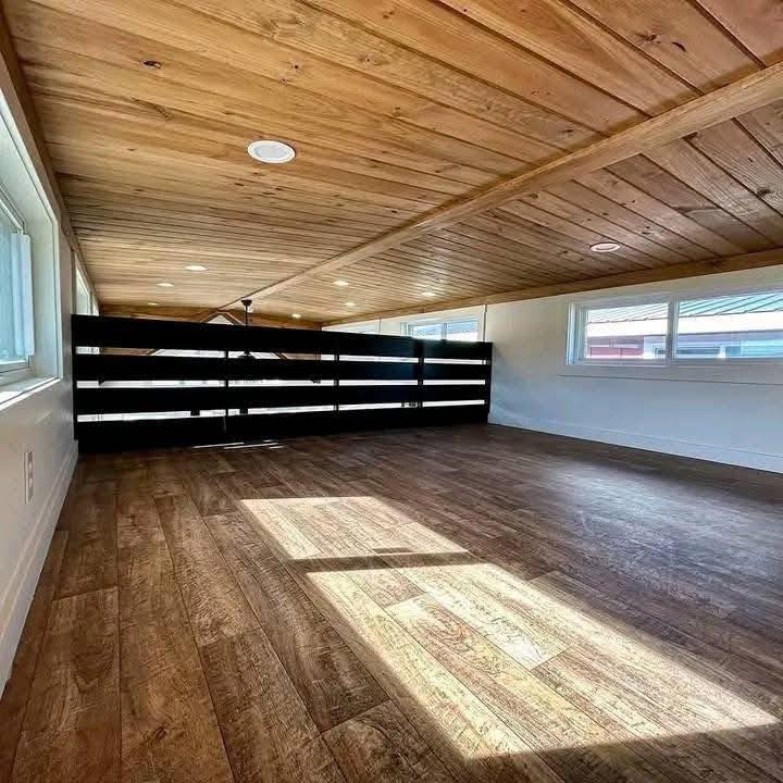 Spacious loft area with wooden floors and ceiling, illuminated by sunlight streaming through windows. A dark wooden railing is visible, creating a cozy ambiance.