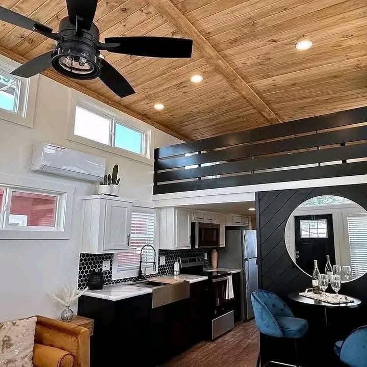 Modern tiny home interior with wood ceiling and loft. Black and white kitchen features a farmhouse sink and hexagonal tile backsplash. Cozy dining nook with round mirror and blue chairs. Ceiling fan and track lighting enhance the warm, stylish ambiance.