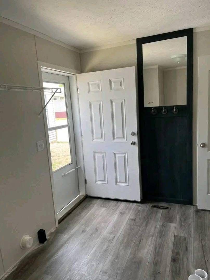 Compact room with light wooden flooring, white walls, and a door. A floor-length mirror with hooks is mounted on a black panel, adding a modern touch.