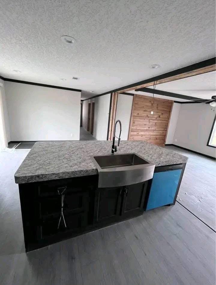 Modern kitchen with a granite island, stainless steel sink, and blue dishwasher. Open layout with wood accents and a calm, minimalist tone.