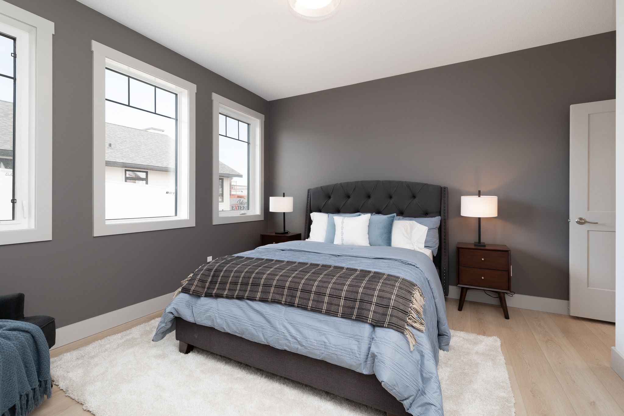 A modern bedroom with gray walls features a bed with a tufted headboard, blue bedding, and a checkered throw. Three windows, two lamps, and a plush rug complete the cozy ambiance.
