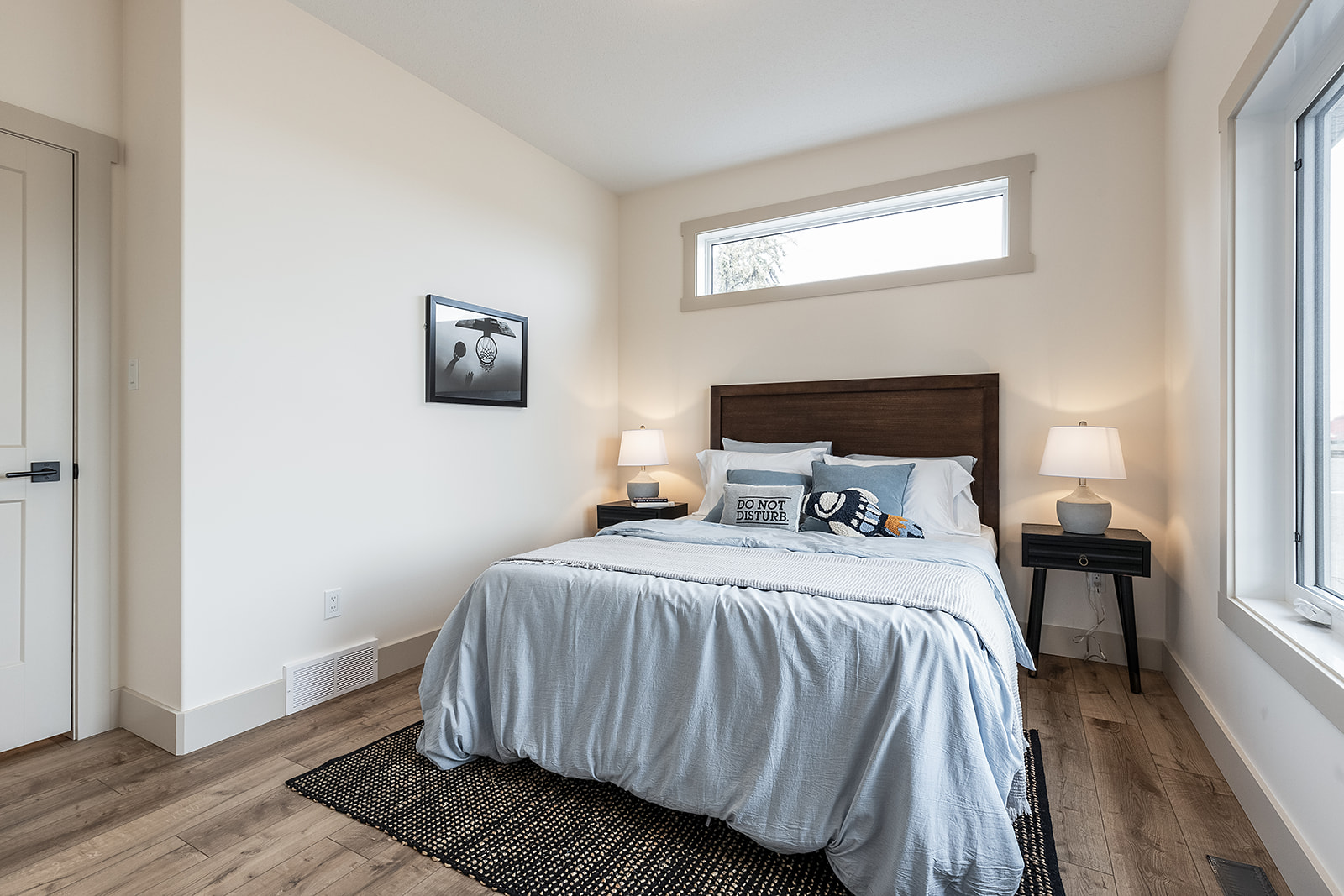 A bright bedroom with a large window, a bed with light blue bedding, pillows including one reading "Do Not Disturb," two side tables, and wall art. Calm ambiance.