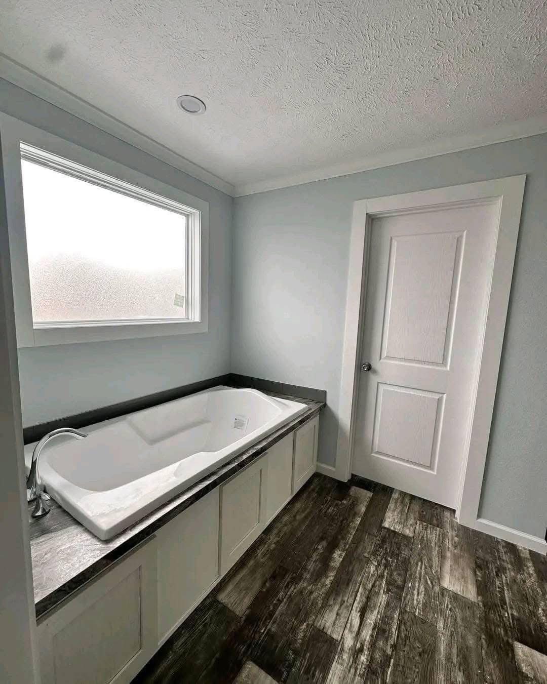 Modern bathroom with a white bathtub, gray countertop, light blue walls, and a frosted window. The wood-textured floor adds warmth.