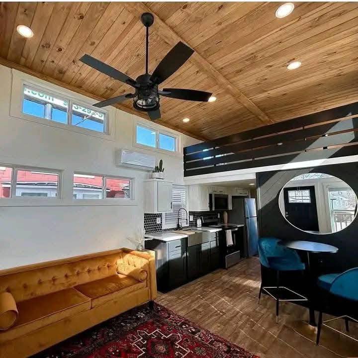 Stylish tiny home interior with wood ceiling, black fan, mustard sofa, patterned rug, sleek black-and-white kitchen, and round mirror above blue chairs.