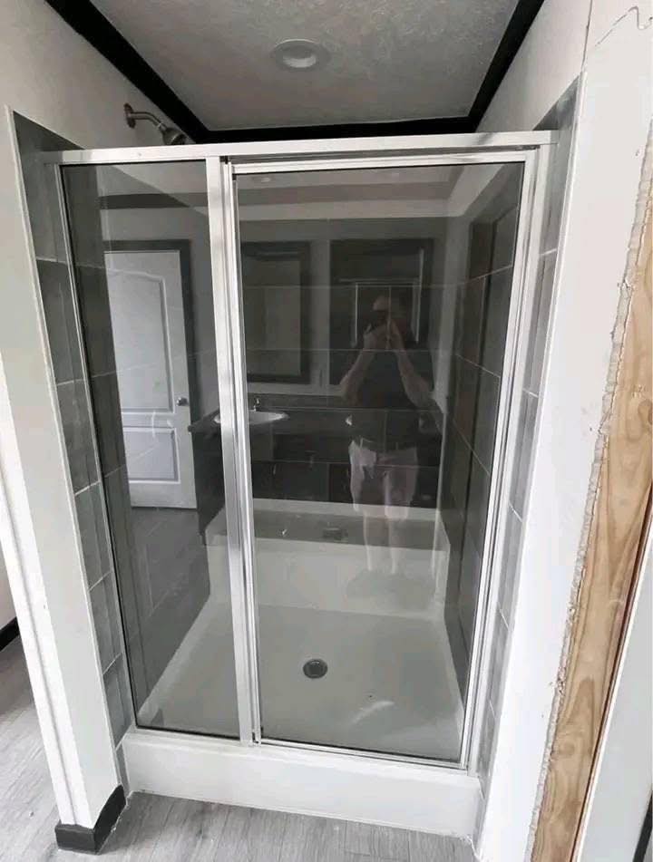 Shower with glass doors facing a mirrored reflection of a person taking a photo. Gray tiles and a wooden trim under renovation. Calm and neutral tone.