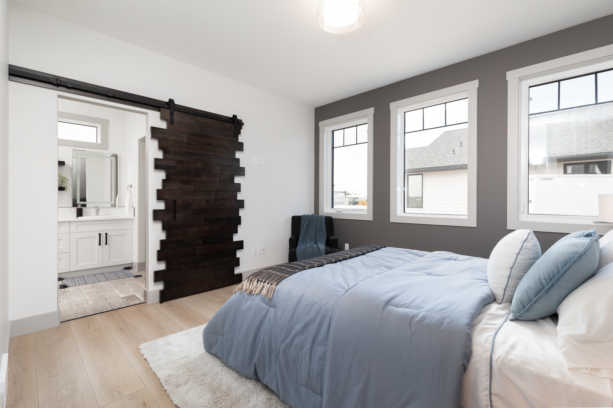 A modern bedroom with a cozy atmosphere features a bed with blue bedding, large windows, a gray accent wall, and a wooden sliding door leading to a bathroom.