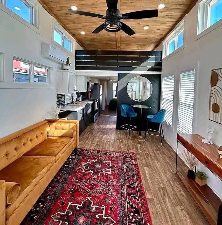 Stylish tiny home with wood ceiling, large fan, mustard sofa, red patterned rug, and cozy dining nook with blue chairs. Bright ambiance.