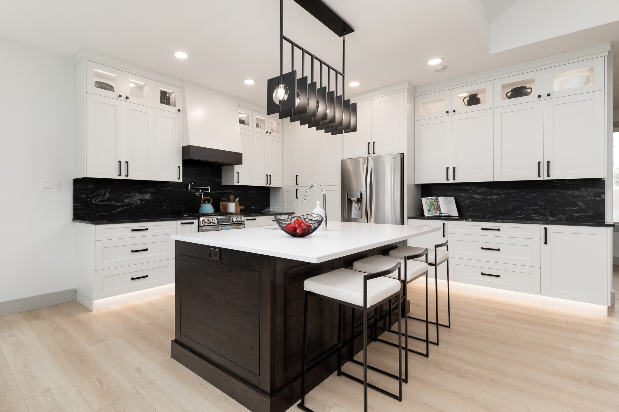 Modern kitchen with white cabinets, black countertops, and a large island. Four barstools, hanging lights, and a stainless steel fridge emphasize elegance.