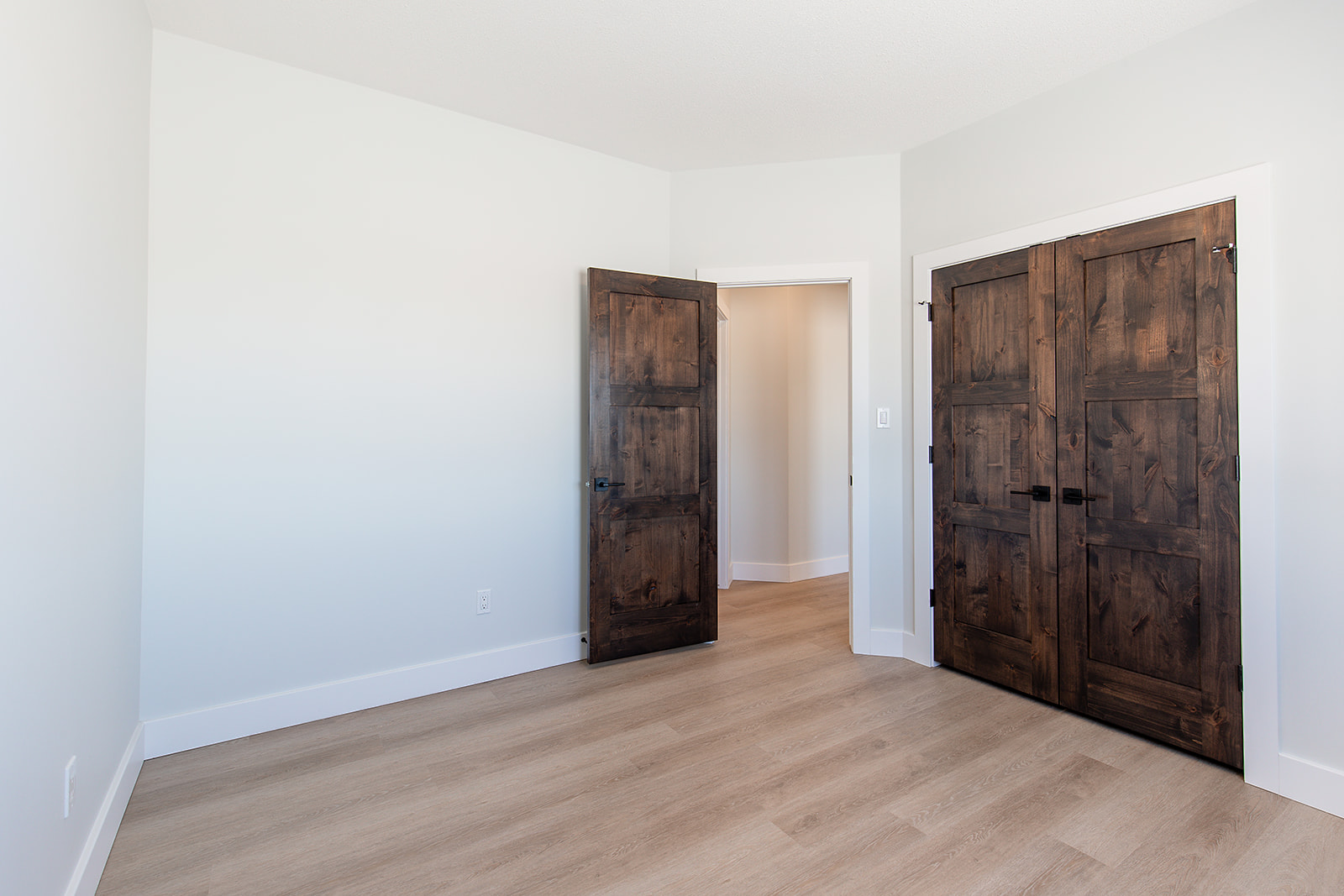 Minimalist empty room with light wood flooring, white walls, and two dark wooden doors. One door is open, revealing a softly lit hallway.