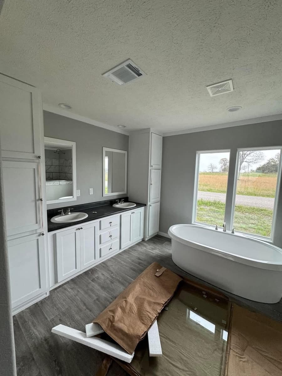Spacious bathroom with a modern design, featuring a freestanding white bathtub by large windows, dual sinks with mirrors, and gray flooring.