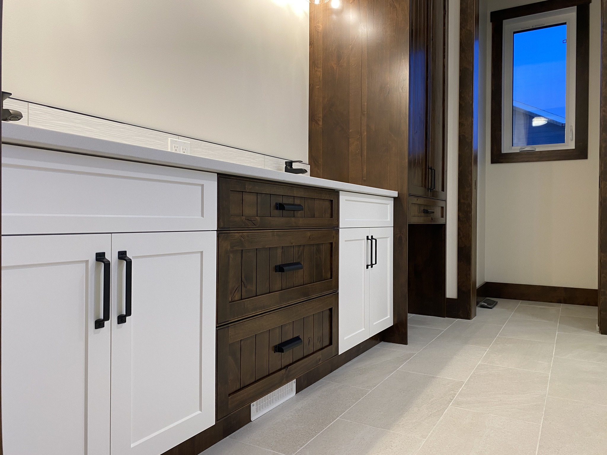 Modern bathroom with white and wood cabinets, black handles, and a light beige tile floor. A small window shows a clear evening sky, adding a calm ambiance.