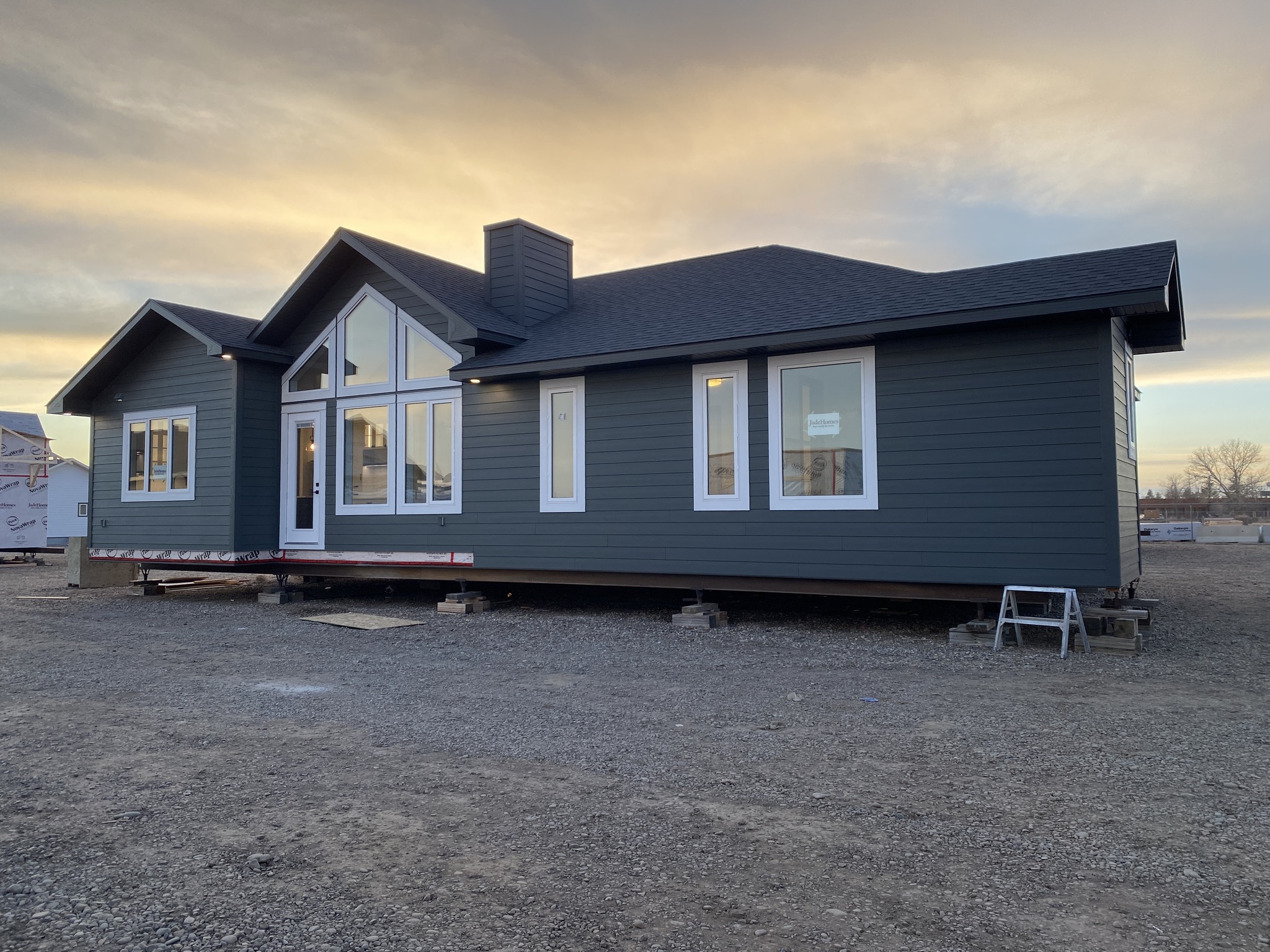 A dark green modular home is elevated on blocks outdoors at sunset. Large windows reflect the golden sky, creating a calm and peaceful atmosphere.
