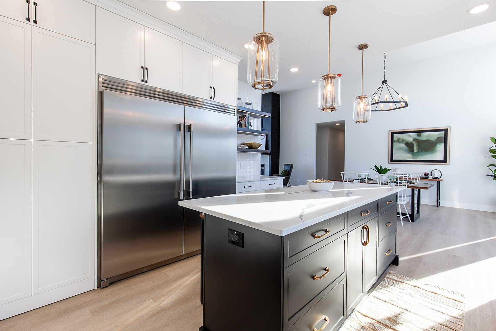 Modern kitchen with a sleek island, pendant lights, and stainless steel fridge. White cabinets, wooden floor, and dining area create a bright, inviting space.