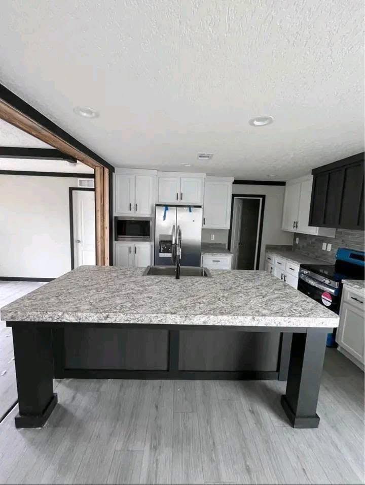 Modern kitchen with a large marble island, stainless steel sink and faucet, surrounded by white cabinets, fridge, and dark countertops. Bright and spacious.
