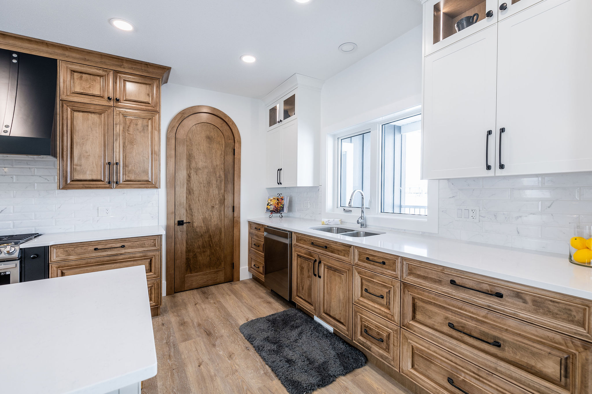 Modern kitchen with wooden cabinets and white countertops, featuring a curved wooden door, stainless steel appliances, and bright natural light.