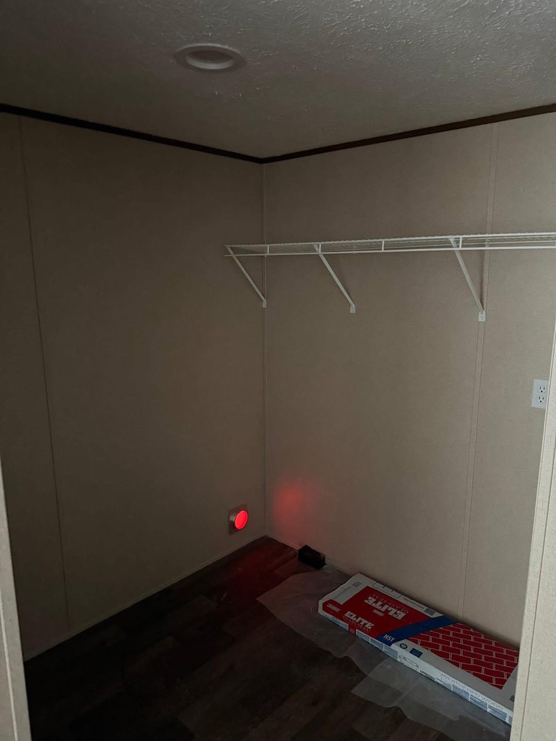 Empty beige room with a wire shelf on the wall. A red glowing light is near the floor, next to HVAC filters and a duct vent. The space feels bare and utilitarian.