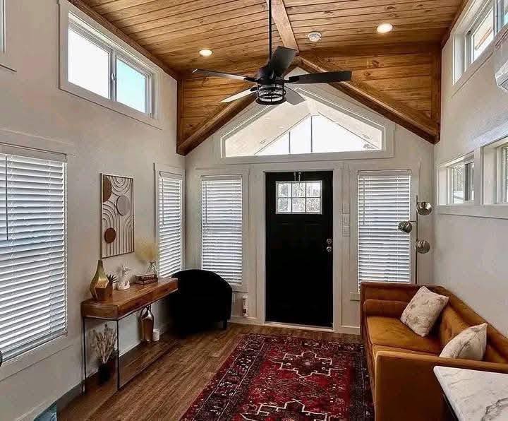Warm, inviting tiny home interior with wooden cathedral ceiling, cozy rug, mustard sofa, and decorative elements. Natural light through large windows.