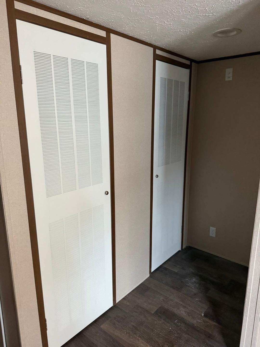 Narrow hallway with dark wood flooring, beige walls, and two white, louvered closet doors with brown trim, creating a simple, cozy atmosphere.