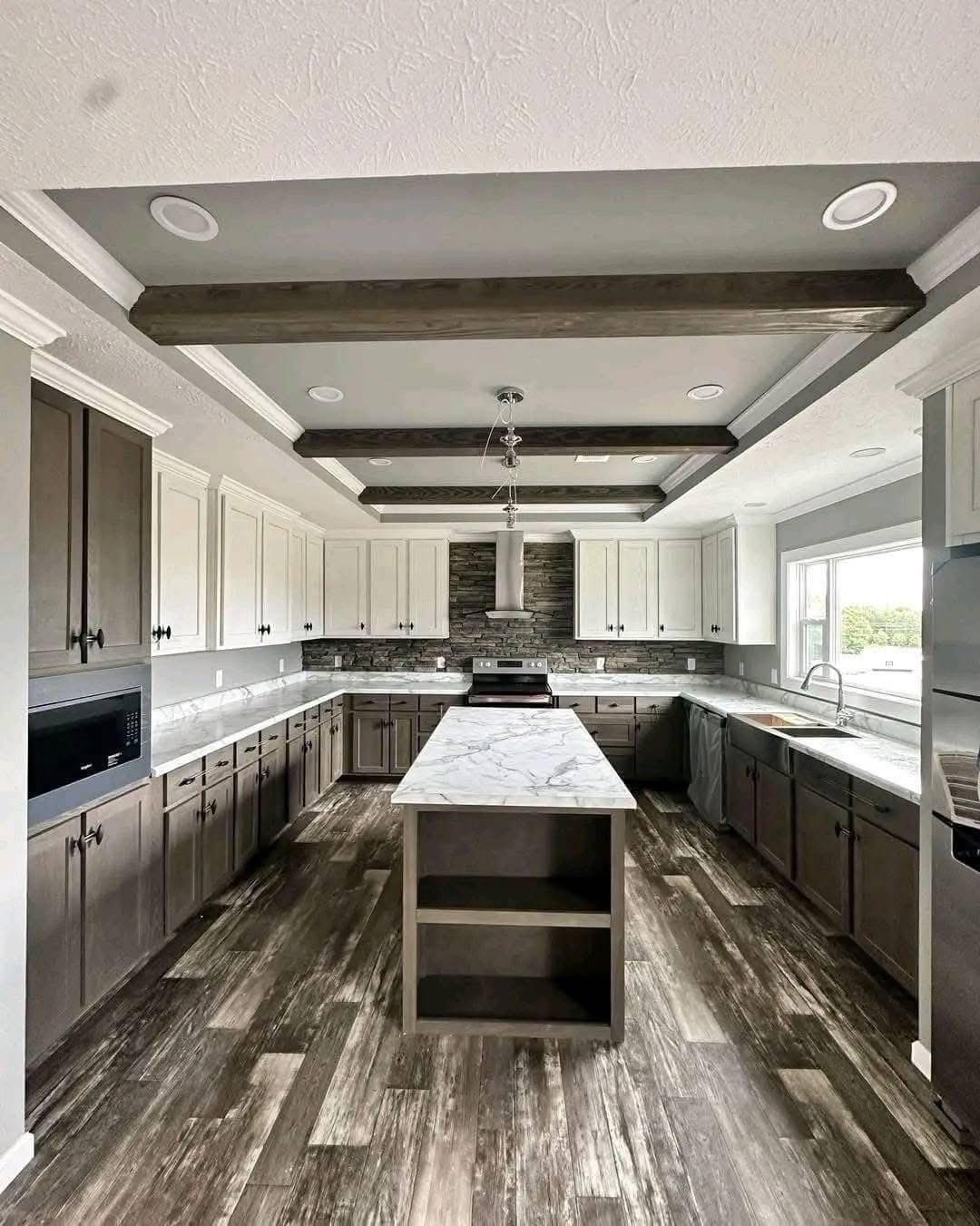 Spacious kitchen with a modern rustic design, featuring a long marble island, dark wood floors, ceiling beams, and a large window providing natural light.