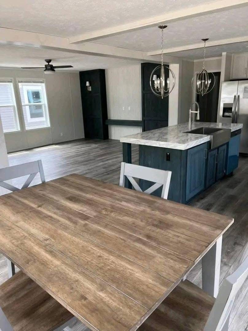 Open-concept kitchen and dining area with a rustic wooden table in the foreground, a blue island with a marble countertop, and two pendant lights.