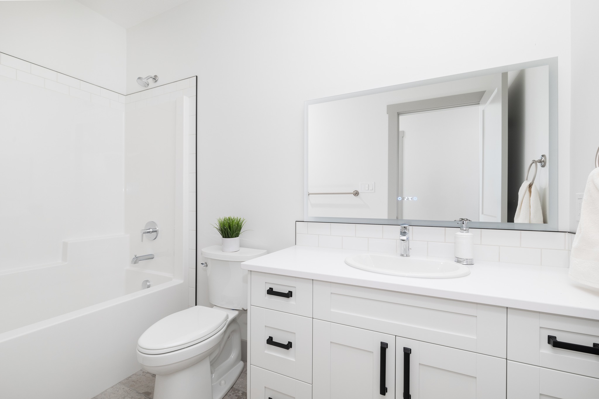 Modern white bathroom with a clean, minimalist design. Features a bathtub-shower combo, toilet, sink on a vanity, large mirror, and a small potted plant.