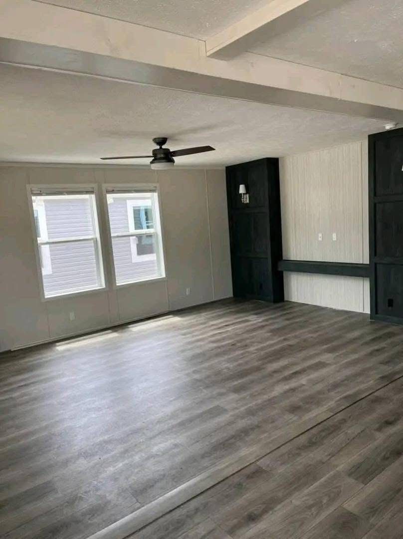 Spacious empty room with light wood flooring, two large windows, a ceiling fan, and built-in dark shelves. Bright and modern interior.