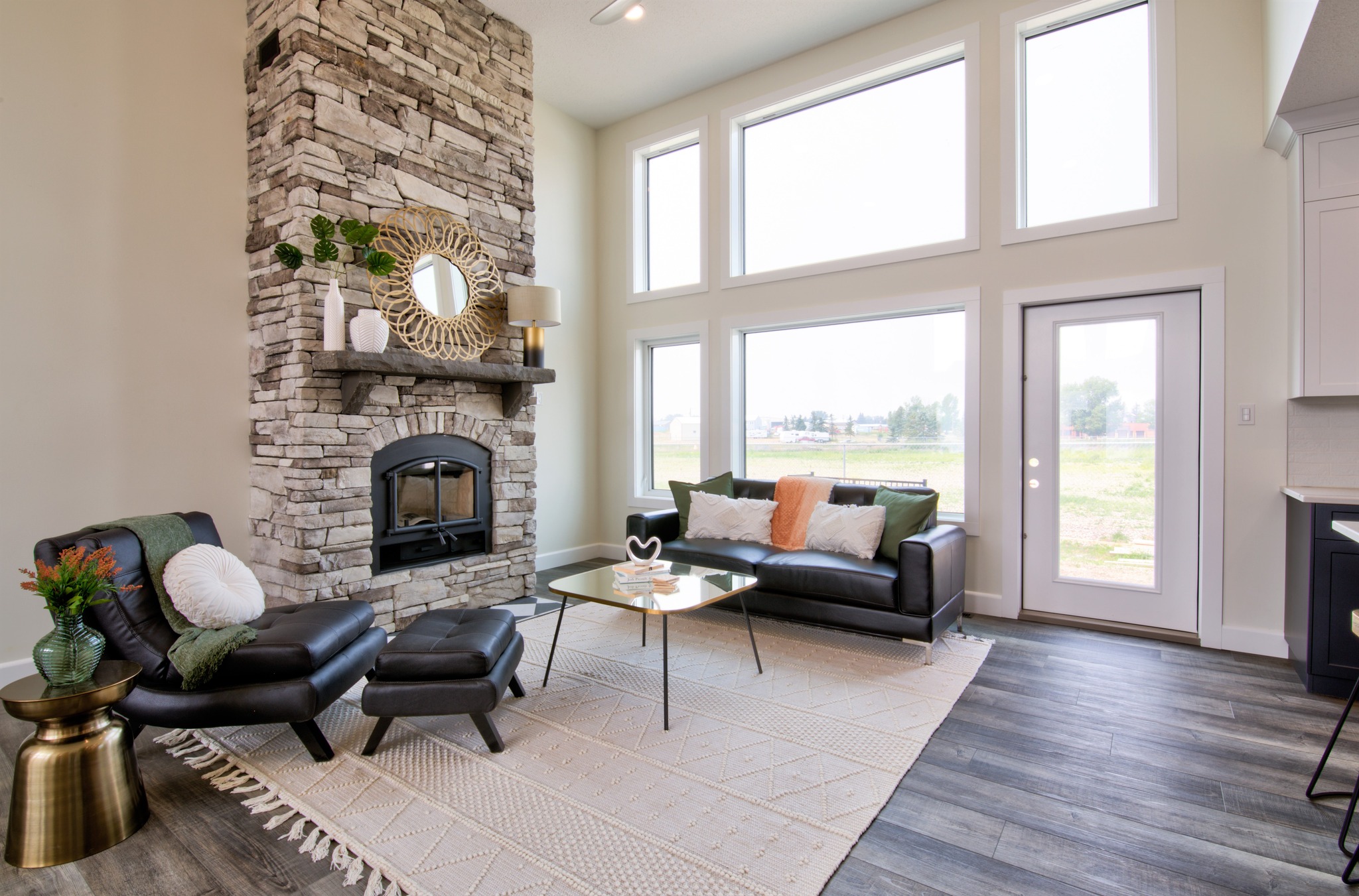 Bright living room with high ceilings, a stone fireplace, and large windows. Features modern furniture, a cozy rug, and decorative plants.