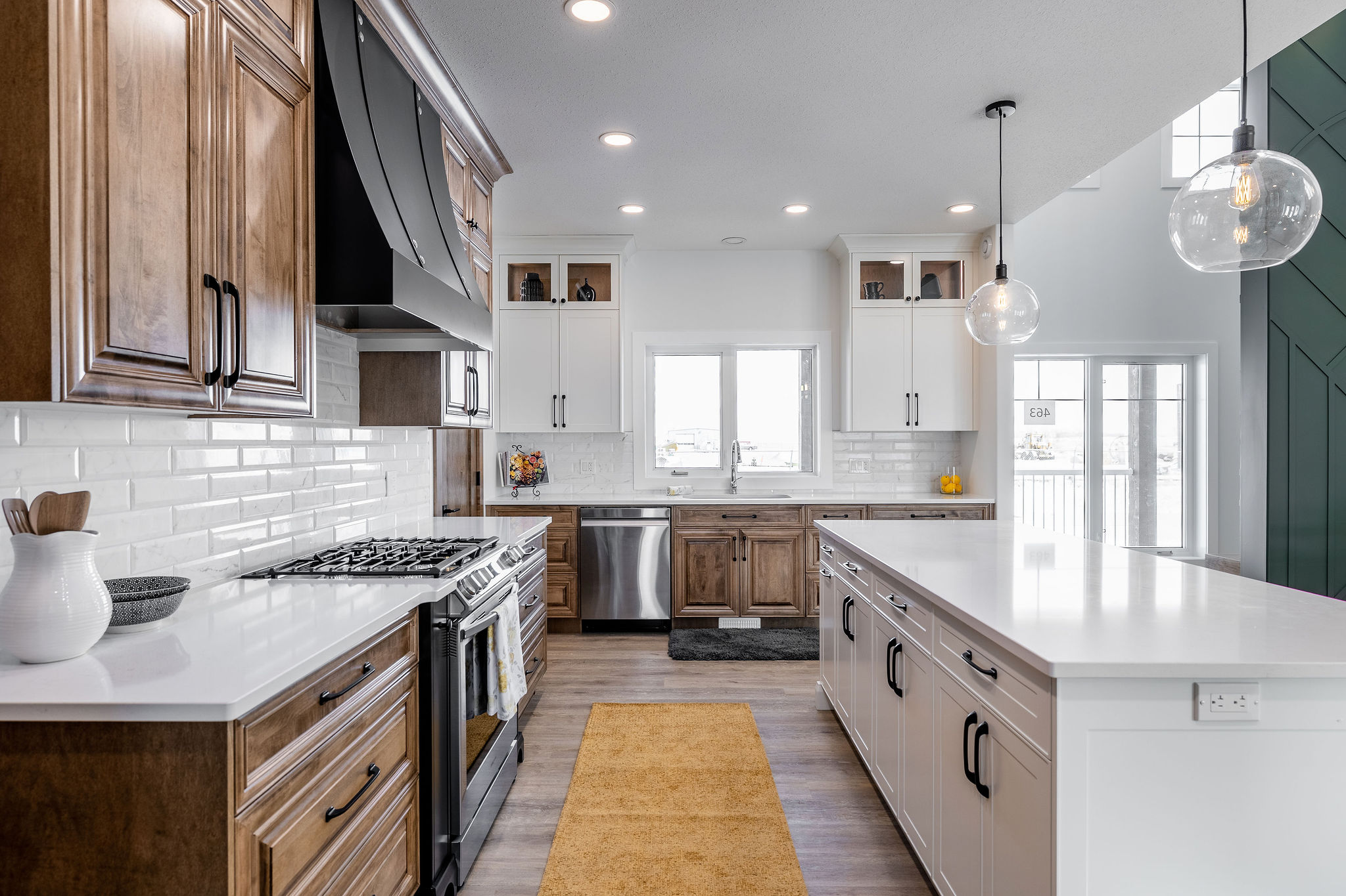 Modern kitchen with wooden cabinets, white countertops, and stainless steel appliances. Bright lighting, large island, and a yellow rug add warmth.