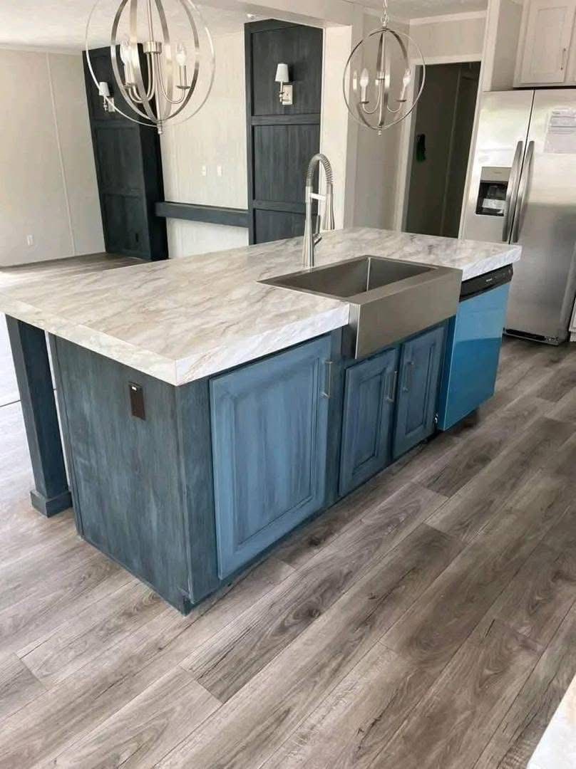 Modern kitchen with a marble-topped island featuring blue cabinets and a stainless-steel sink. Gray wooden floors and chrome light fixtures add elegance.