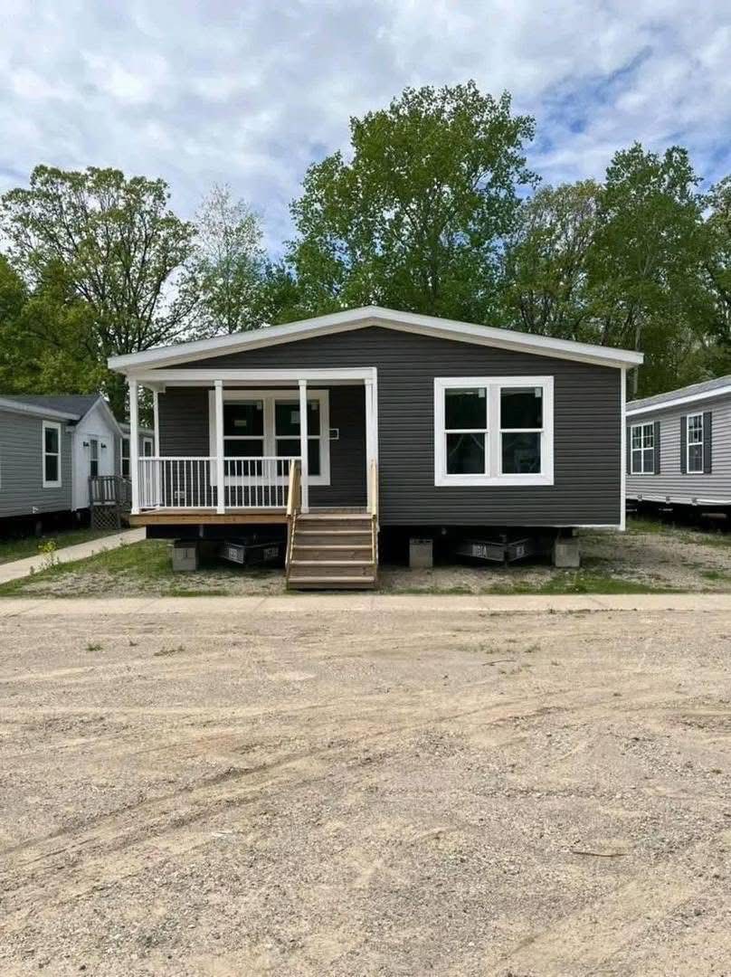 A gray mobile home with a white railing porch sits on a dirt lot, surrounded by trees and a cloudy sky. The scene feels calm and residential.