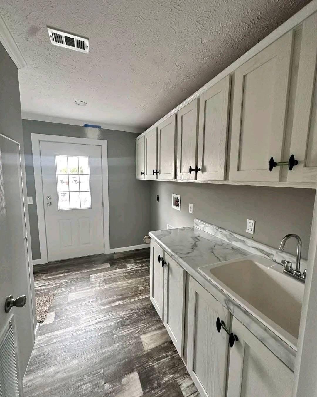 A laundry room with a white sink and marble countertop on the right, light gray wood cabinets above, and a door with a window on the far wall. The flooring is gray wood, creating a modern and clean look.