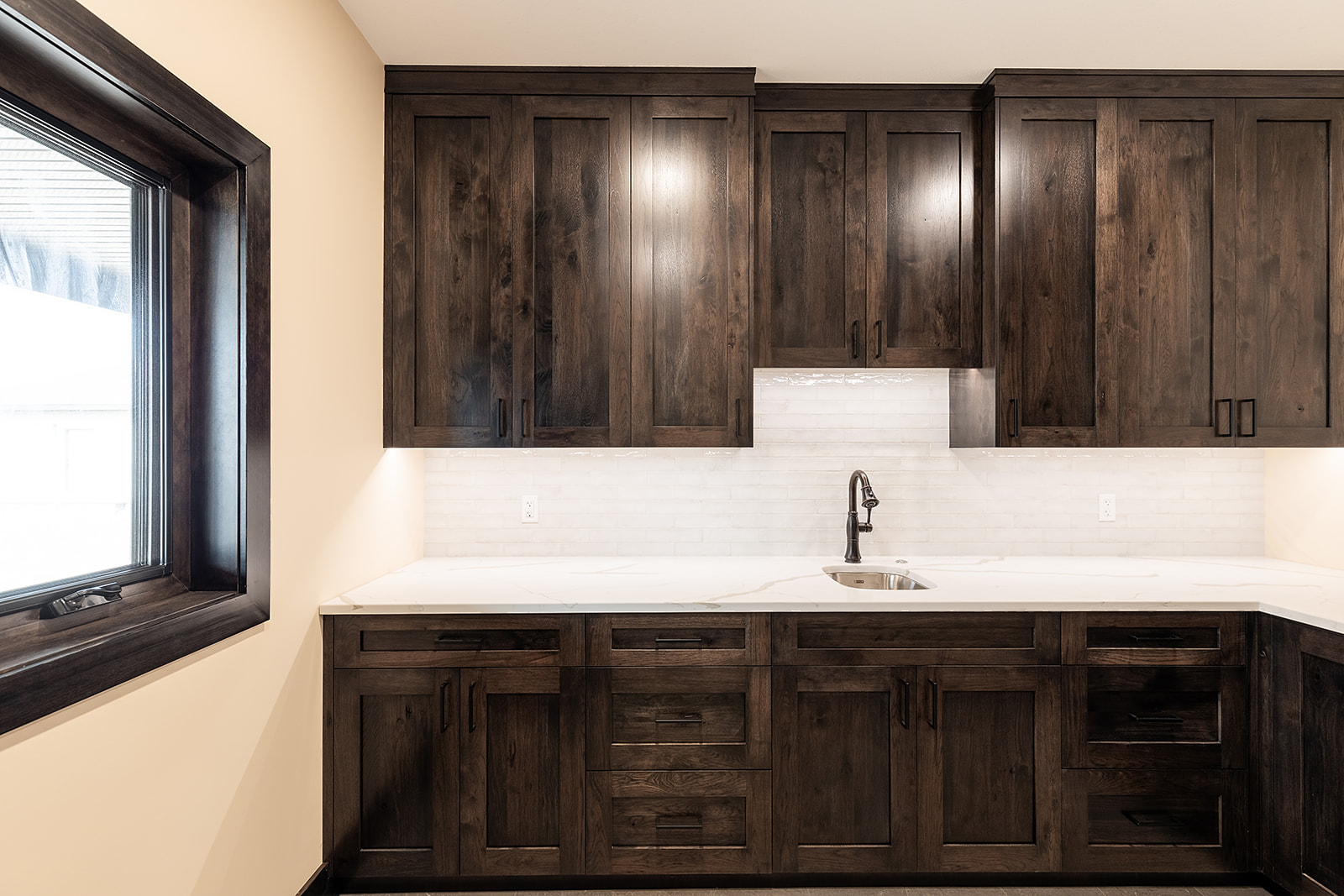 Modern kitchen with dark wooden cabinets, a white backsplash, and a sleek black faucet on a light countertop. The room is brightened by natural light.