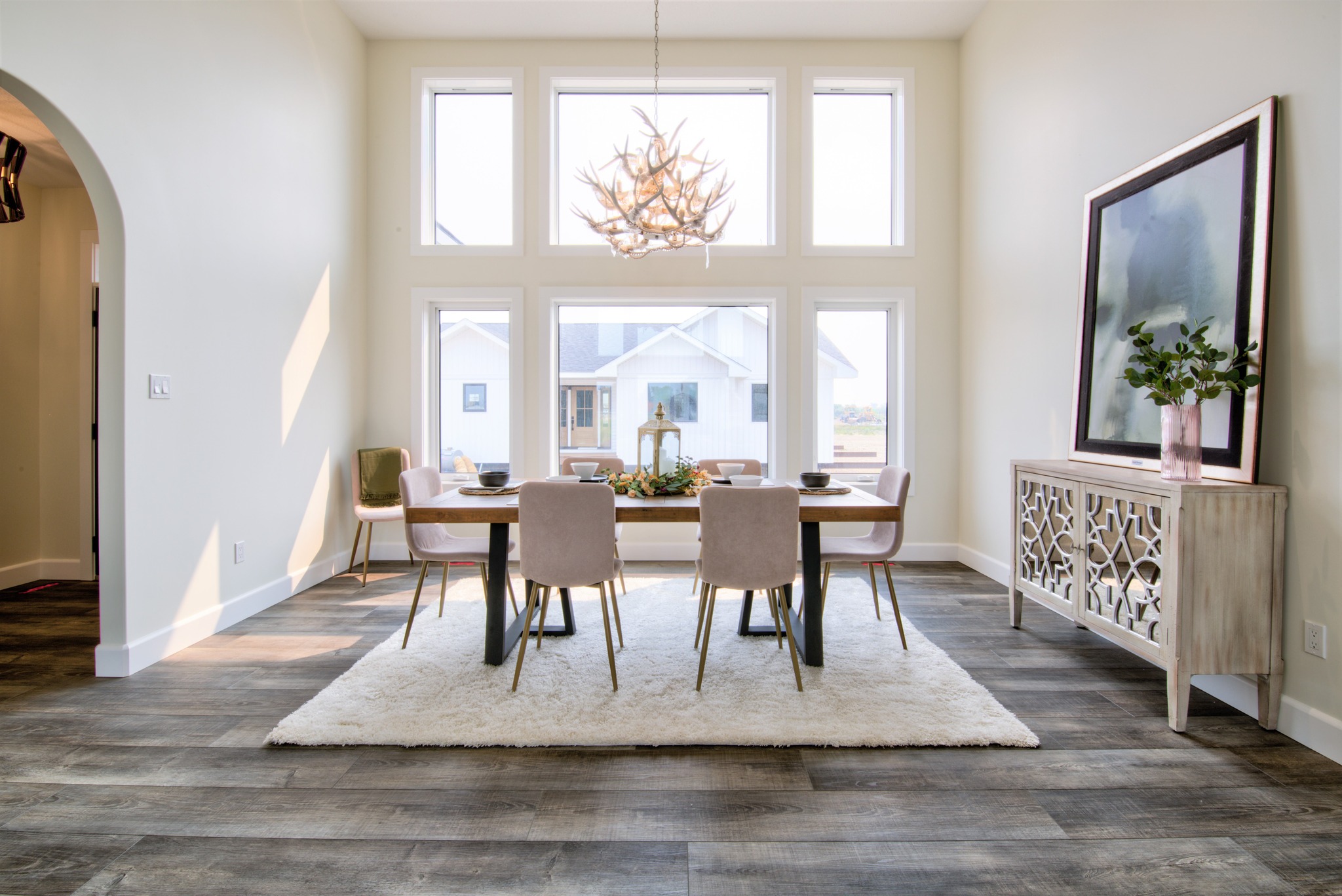 Elegant dining room with large windows, antler chandelier, and modern table with six chairs on a plush rug. Neutral tones, cozy ambiance.