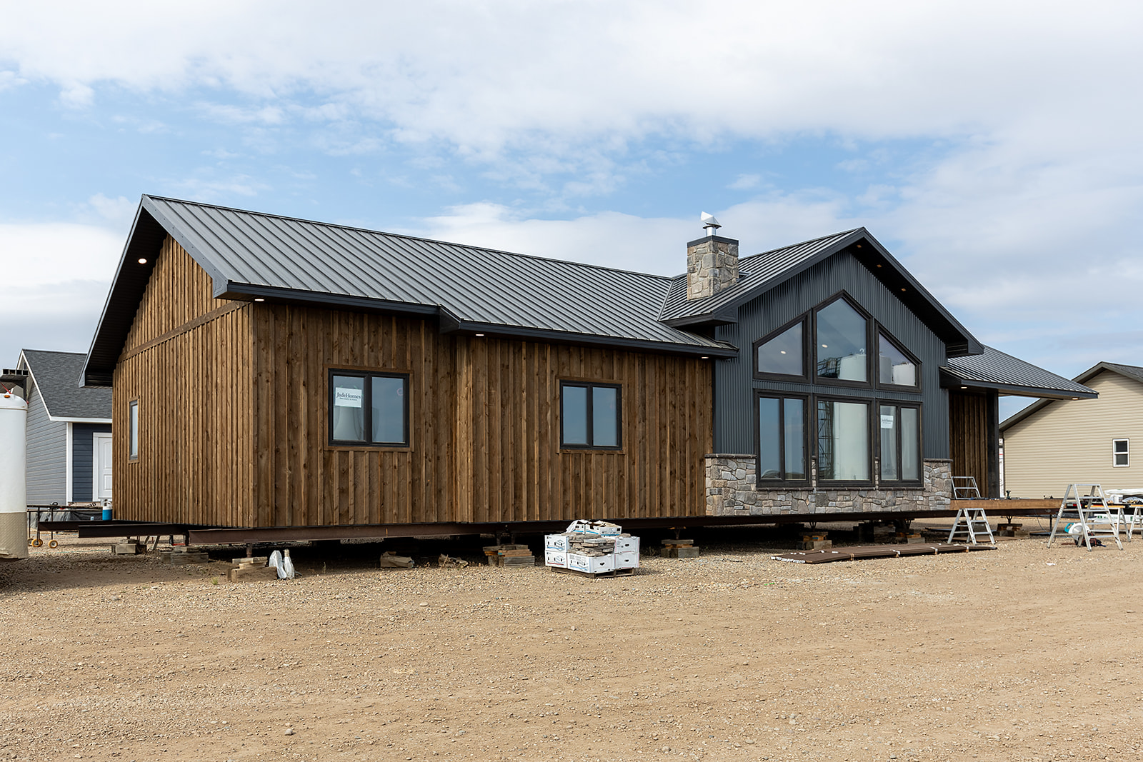 A wooden house with a steep metal roof and large windows sits elevated on a construction site. The ground is bare, with construction materials around.