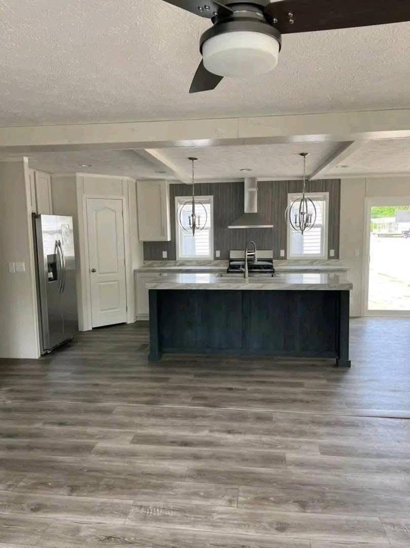 Modern kitchen with gray wood floor, dark island, marble countertop, two pendant lights, stainless steel appliances, and a ceiling fan. Bright and airy.