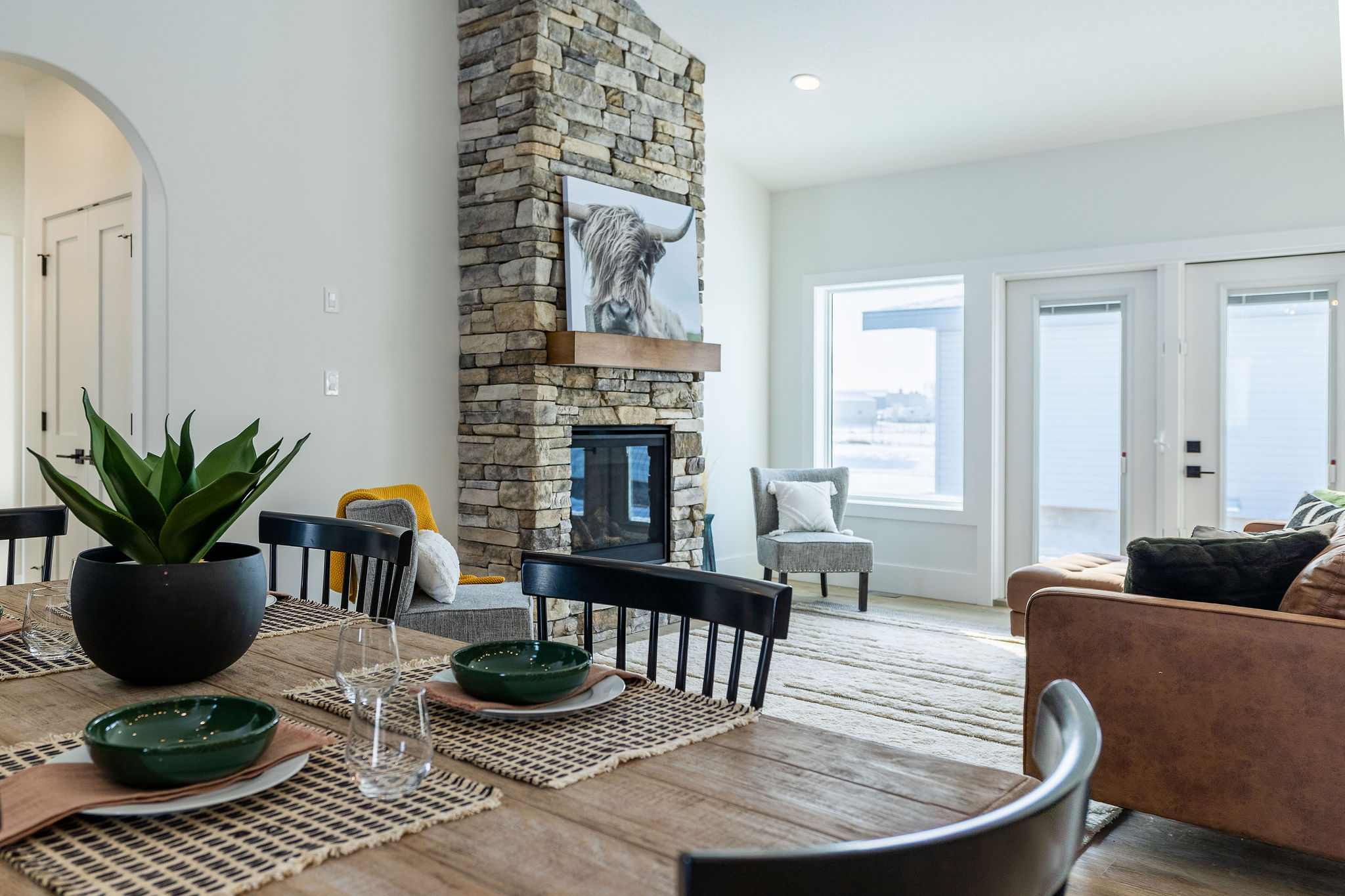 A modern living space features a stone fireplace with a bull painting, a rustic dining table with green dishware, a brown sofa, and large windows, creating a cozy and inviting atmosphere.