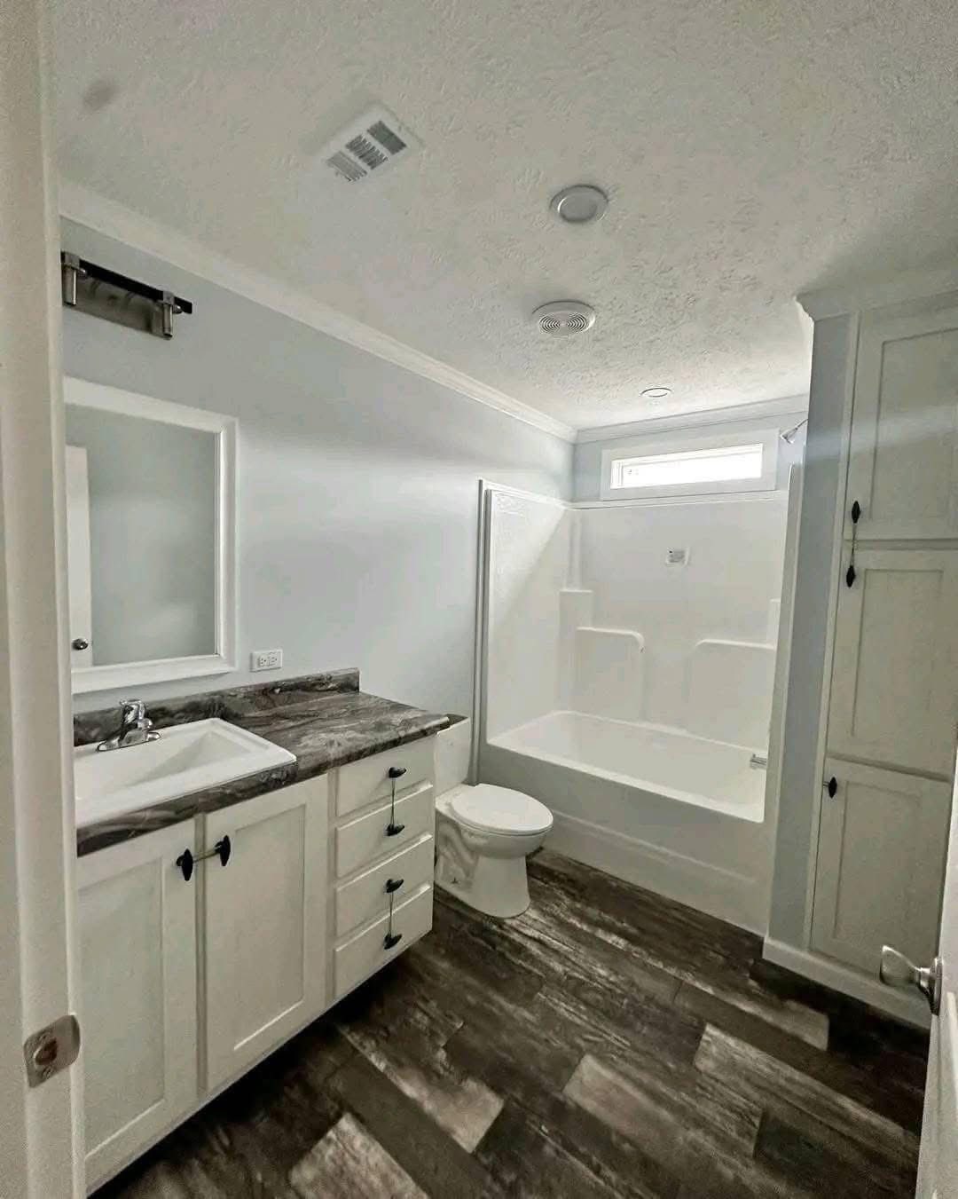 Modern bathroom with white cabinets, marble countertops, and dark wood floors. Features a bathtub, shower, toilet, and a rectangular window.