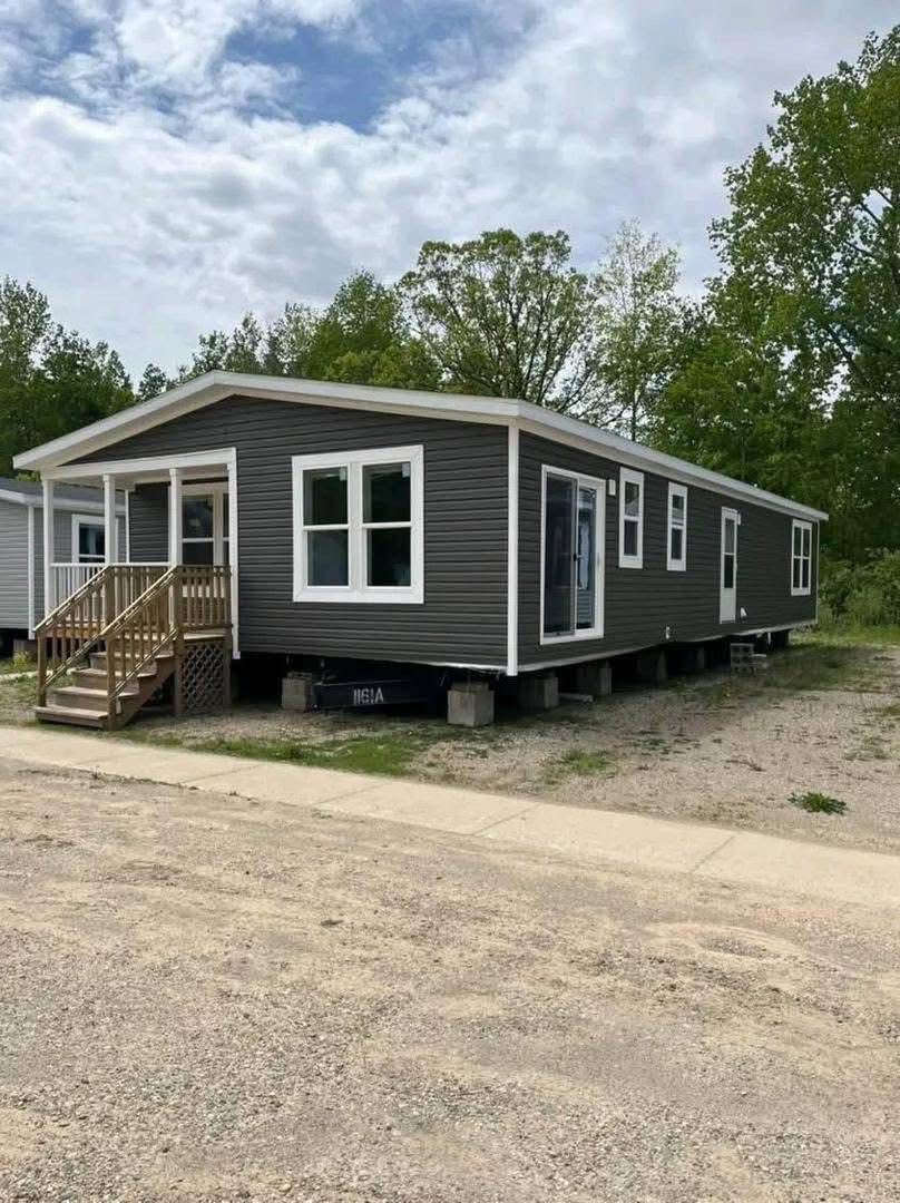 A gray manufactured home with a small wooden porch sits on a gravel lot. Surrounded by trees under a cloudy sky, the scene is calm and spacious.