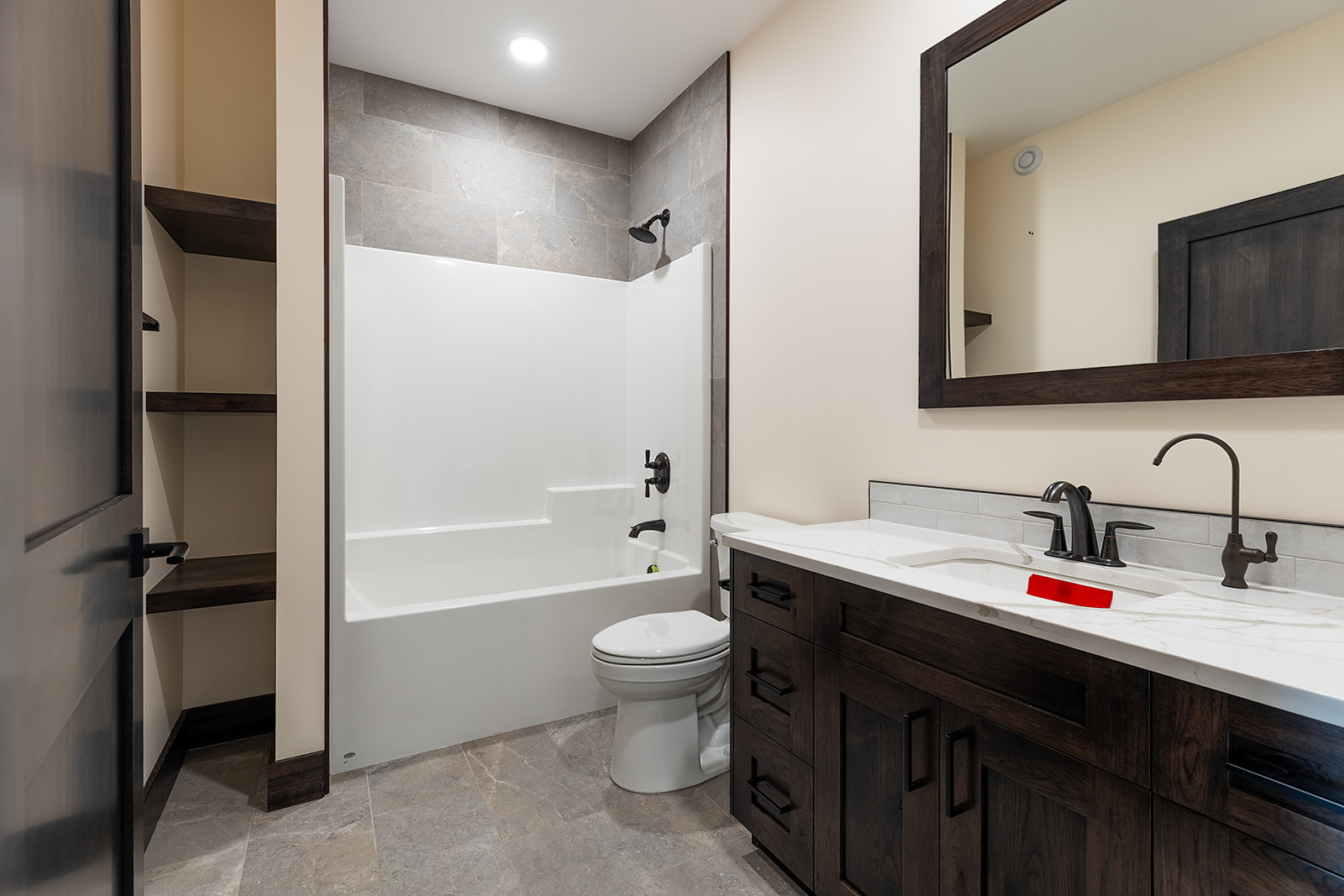 Modern bathroom with beige walls, featuring a white bathtub and toilet. Dark wood vanity with dual sinks, large mirror, and open shelving. Cozy ambiance.
